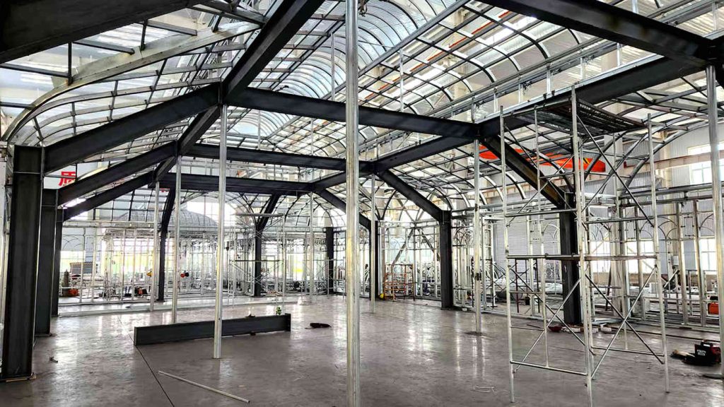 Interior factory view of a large steel-frame sunroom under assembly, showing black structural beams, internal support columns, clear arched roof panels, and construction scaffolding on the right.
